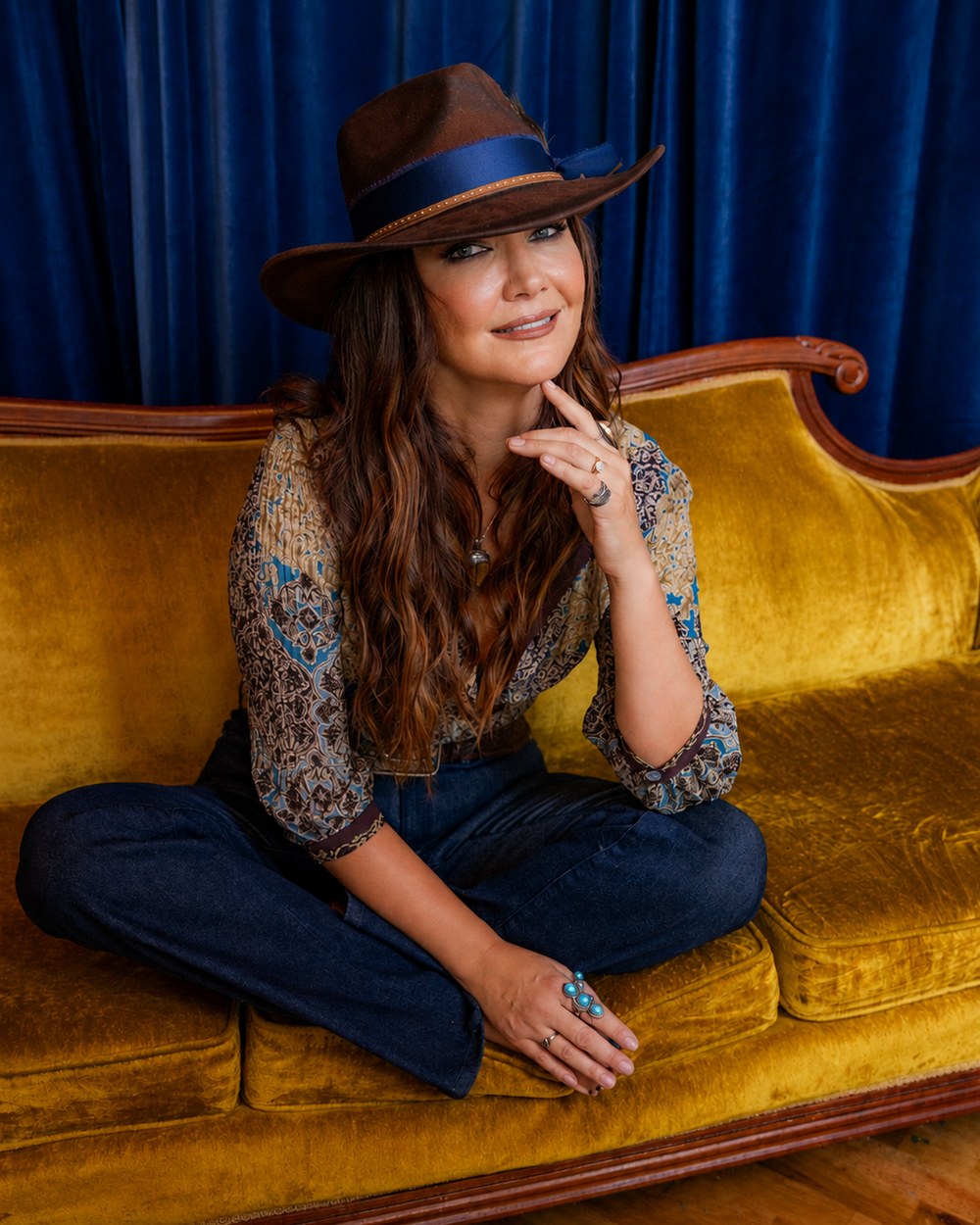 Kris Bradley seated on a gold velvet sofa in a wide-brim hat.
